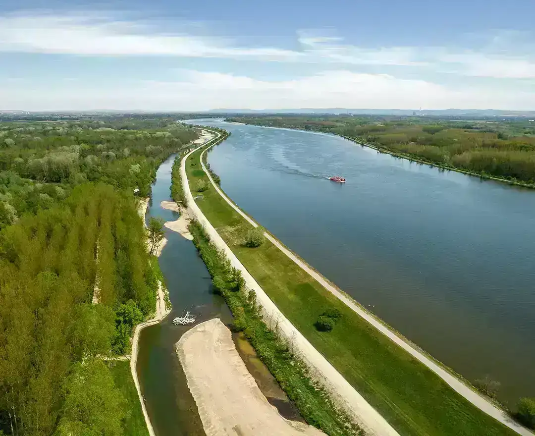 Ein Drohnenflug über die Donau bei strahlendem Sonnenschein. Ein rötliches Schiff bahnt sich seinen Weg auf der Donau. Die entstehende Fischwanderhilfe ist auf der linken Seite zu sehen.